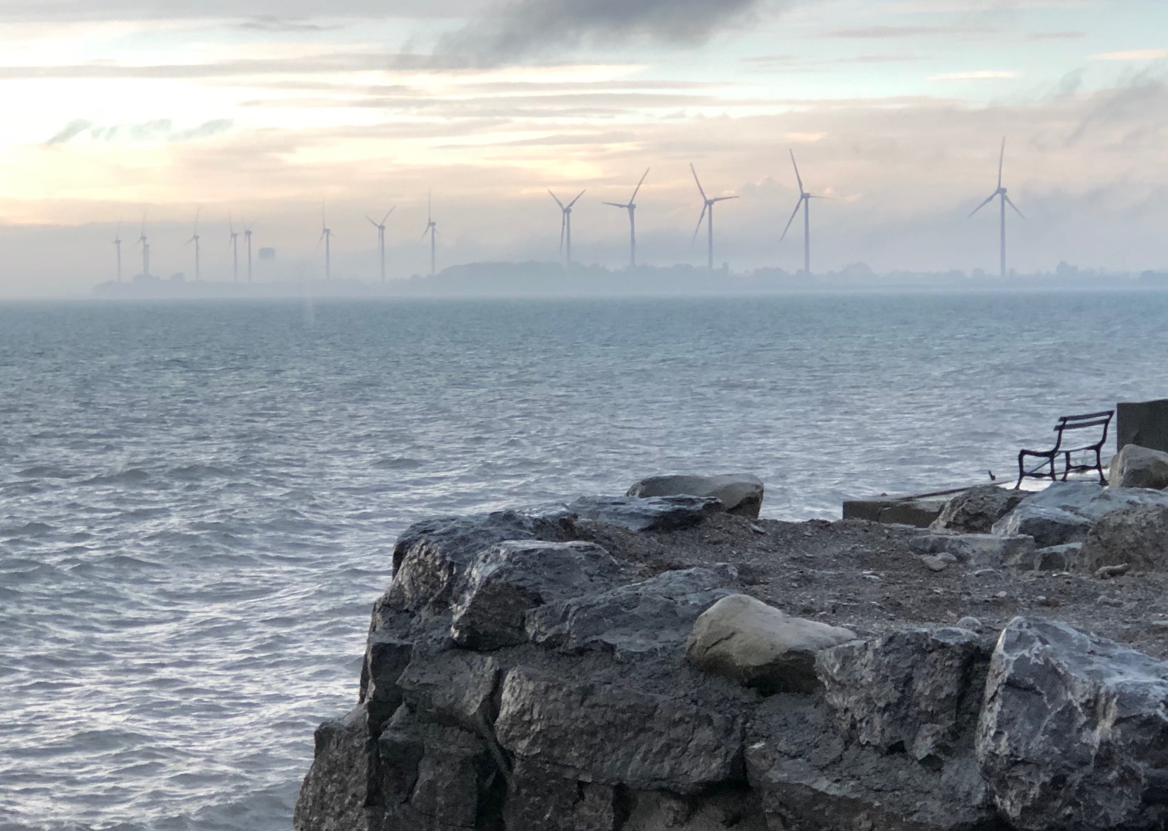 Multiple wind turbines can be seen over a body of water through the fog. A bench is on a stone wall in the foreground.
