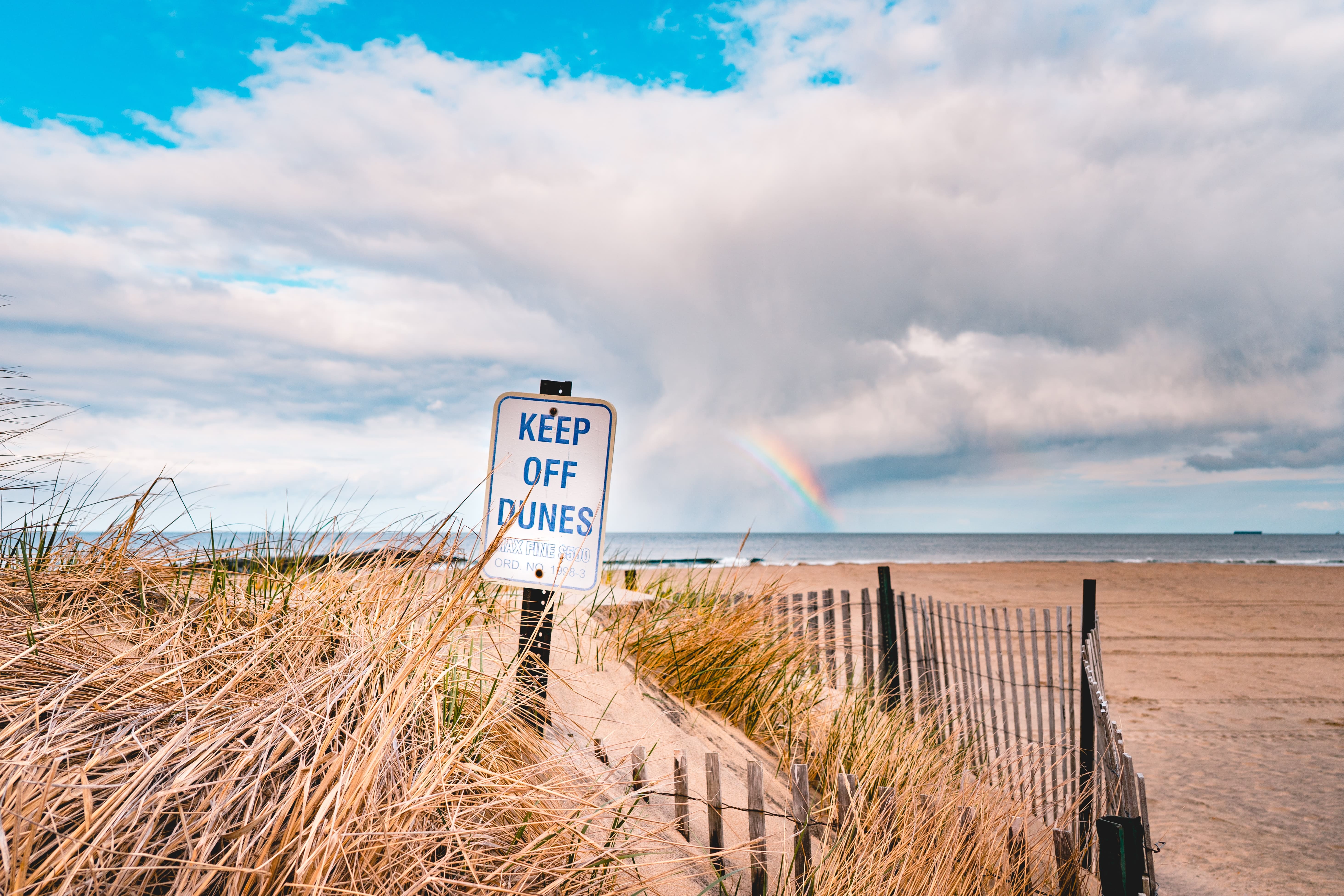 A sign with “Keep off the Dunes” is stuck in a dune with a beach in the background.