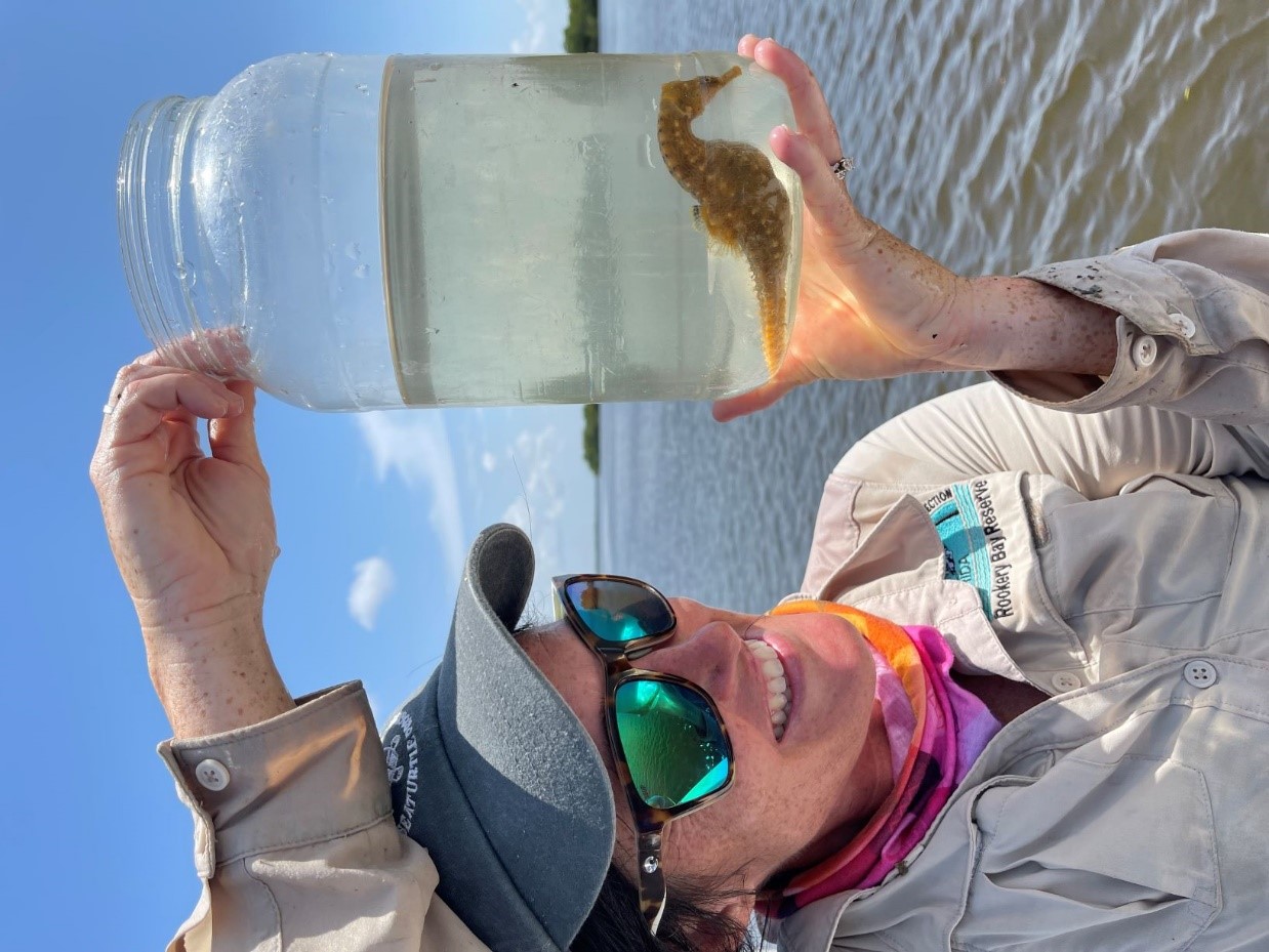 Person smiling while looking at a jar that contains a large seahorse. A body of water is in the background.