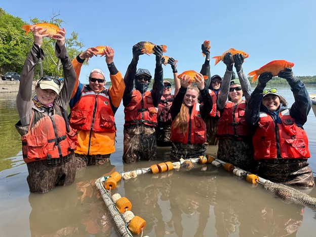 Eight people wearing chest waders holding fish above their heads.