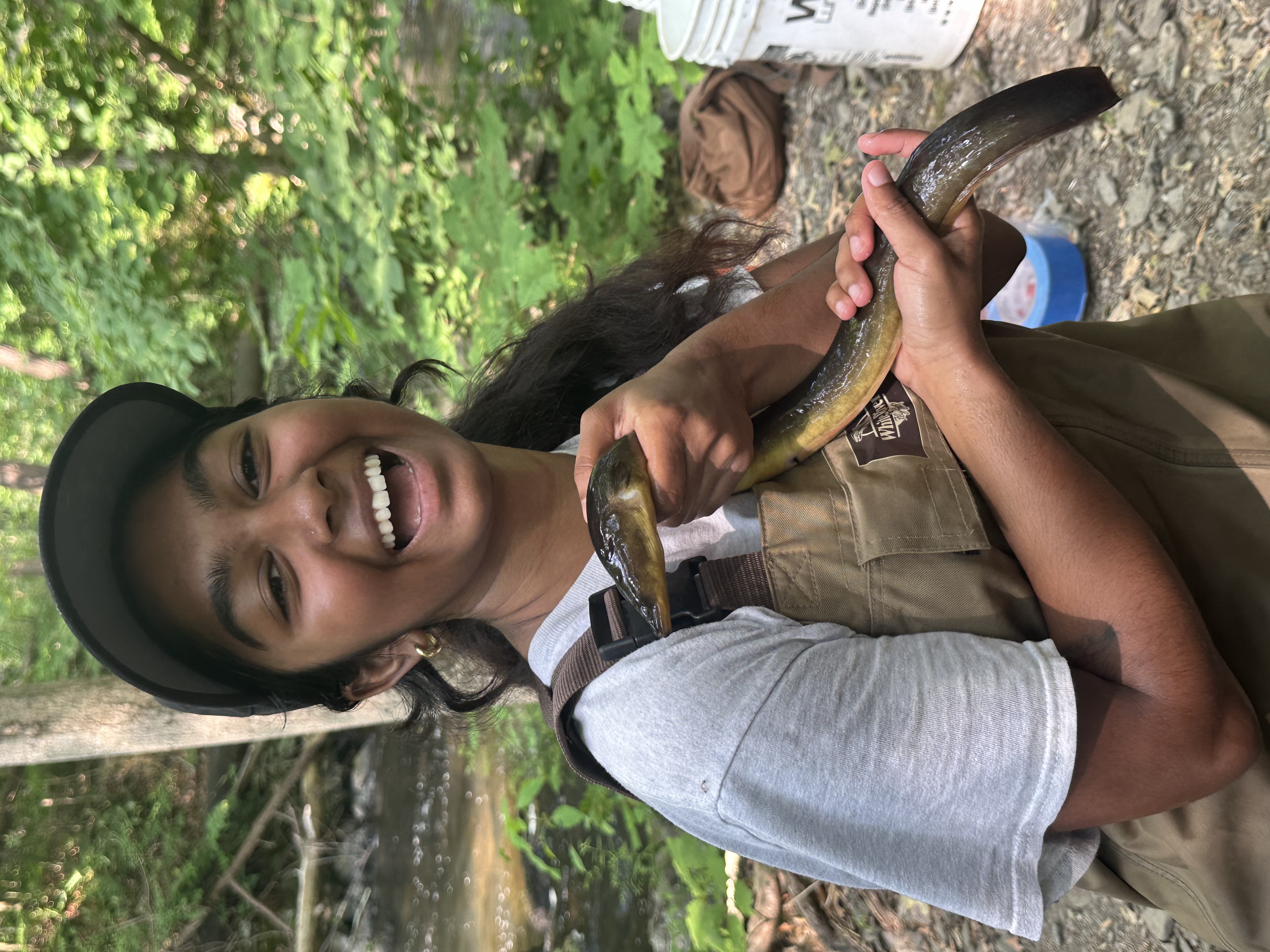 A person outdoors holds an eel with both hands and smiles at the camera. They are wearing overalls and a cap.