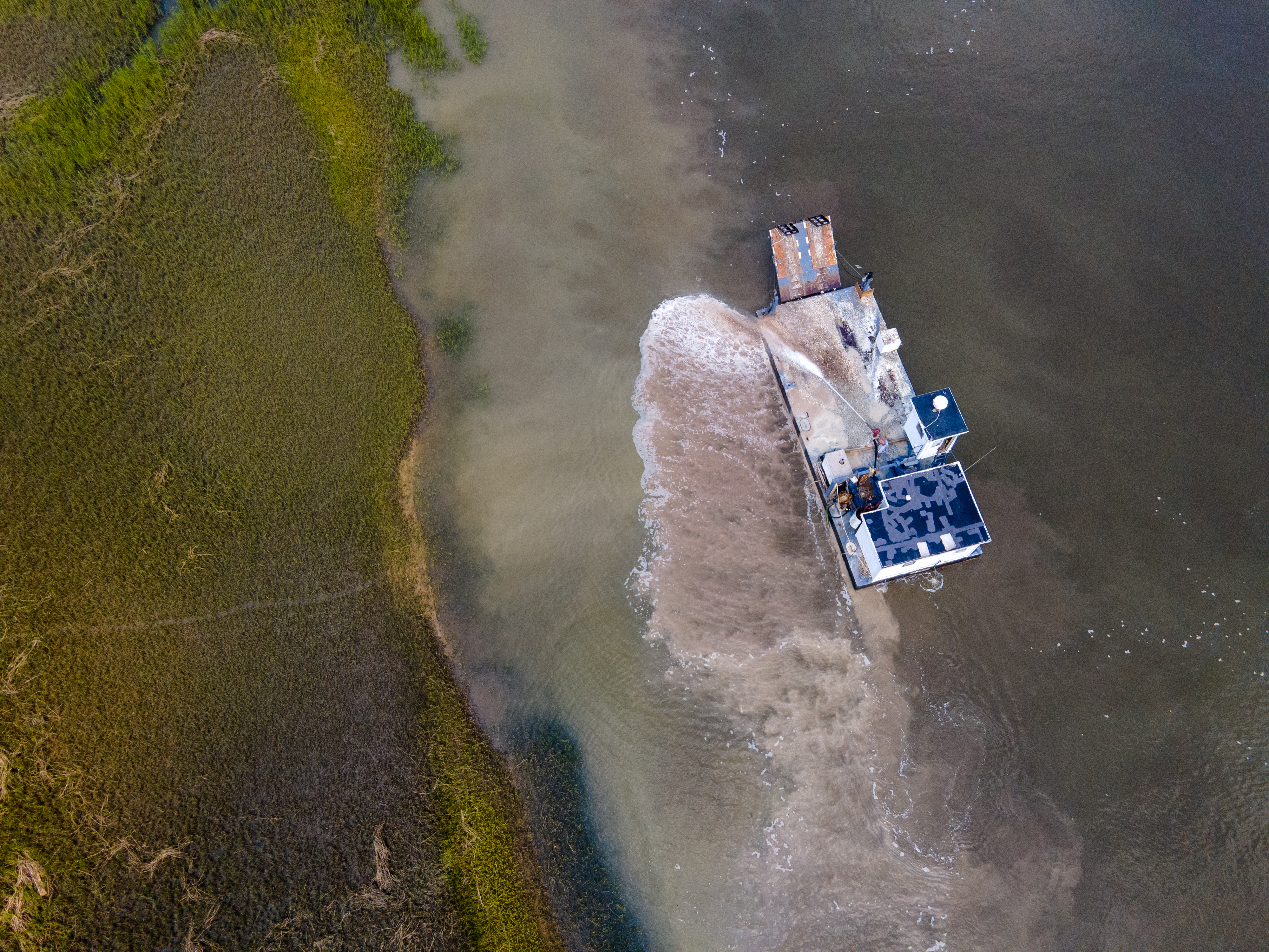 Aerial view of a boat navigating through murky, shallow waters near a grassy wetland. The boat creates a wake as it moves, stirring up sediment.