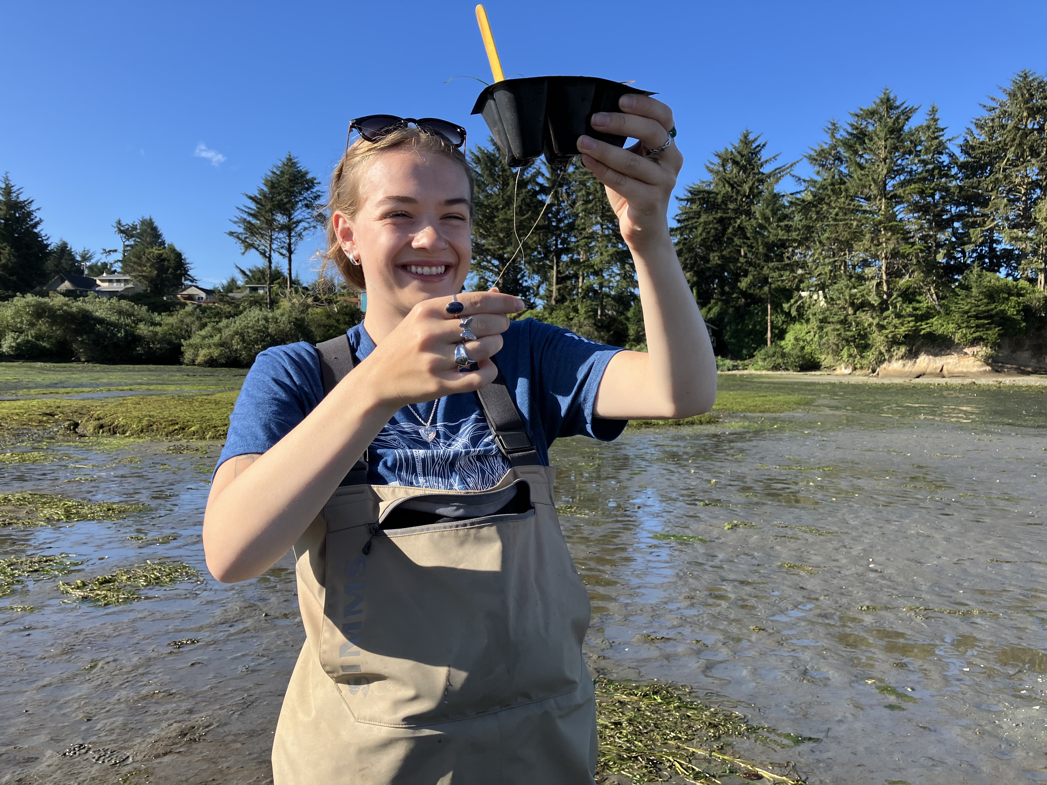 A person in waders stands in a shallow body of water, holding up a black container with a cheerful expression. Trees and greenery are visible in the background under a clear blue sky.