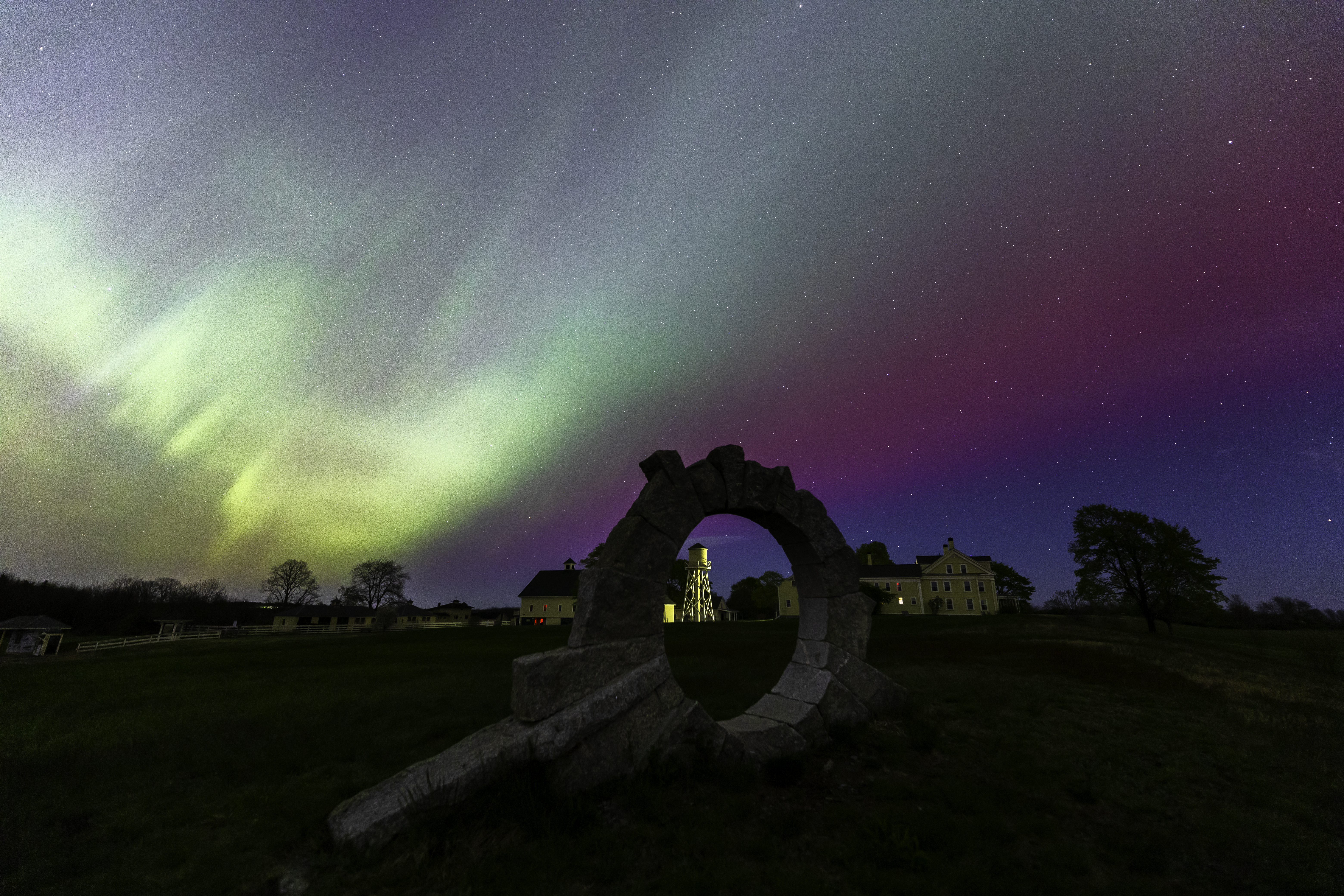 A night sky features vibrant green and purple auroras above a landscape with an ancient stone circle in the foreground. In the background, historic buildings and a few trees are silhouetted against the colorful display of lights.