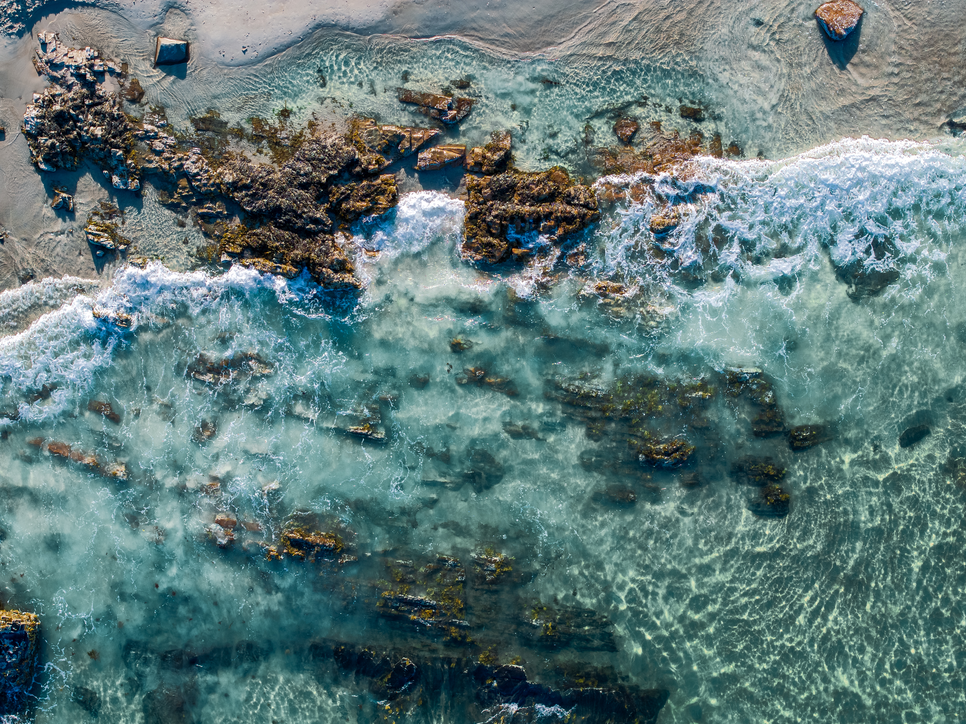 An aerial view of a coastal scene with clear, turquoise water washing over rocky formations and sandy areas.