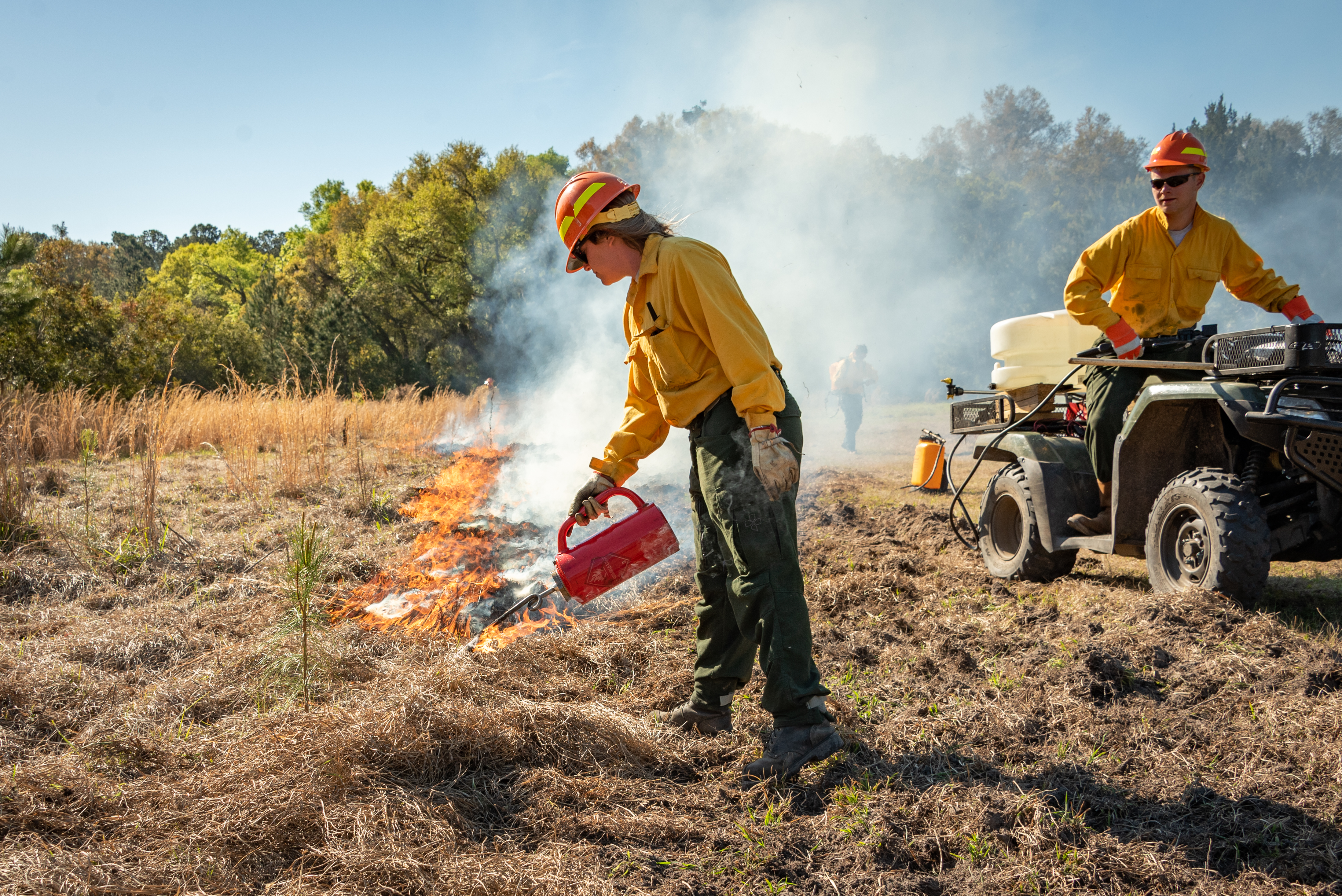 A firefighter in yellow gear uses a drip torch to ignite a controlled burn in a grassy area, while another firefighter stands by an ATV carrying equipment.