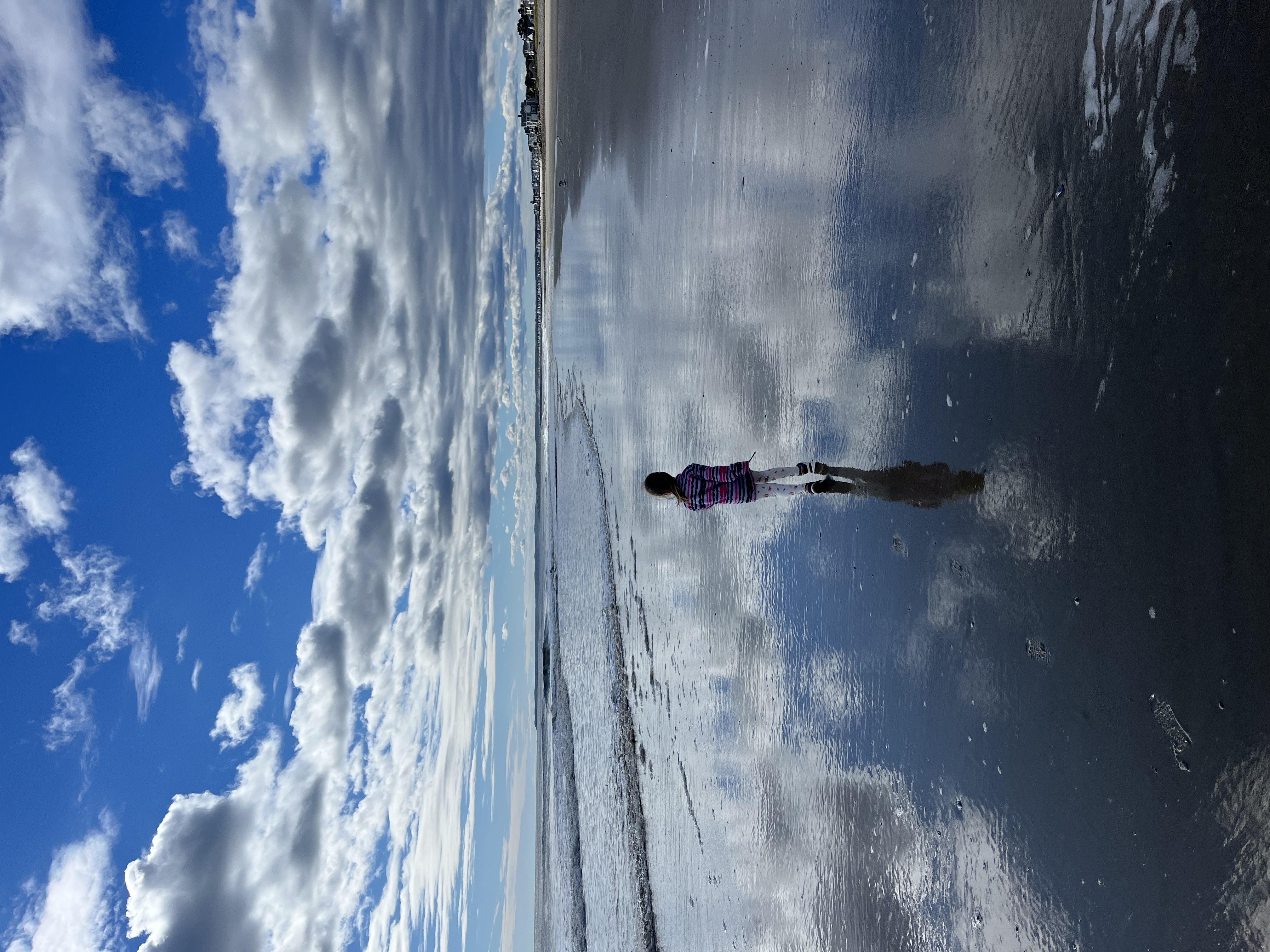 A small child walks on a vast, empty beach with water reflecting the blue sky and scattered clouds.