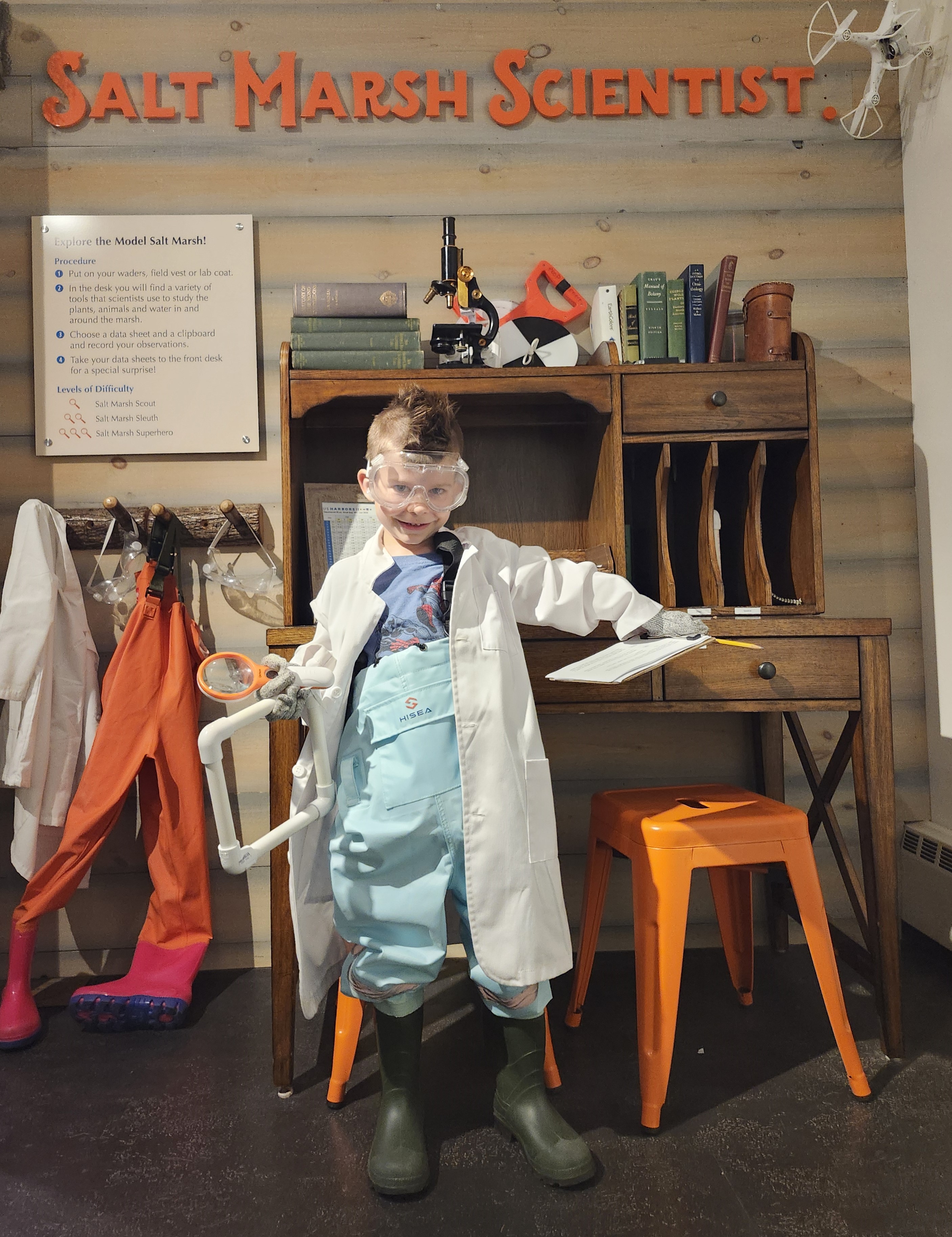 A child dressed in a lab coat, goggles, and green boots stands in front of a wooden desk, holding a clipboard and test tube.