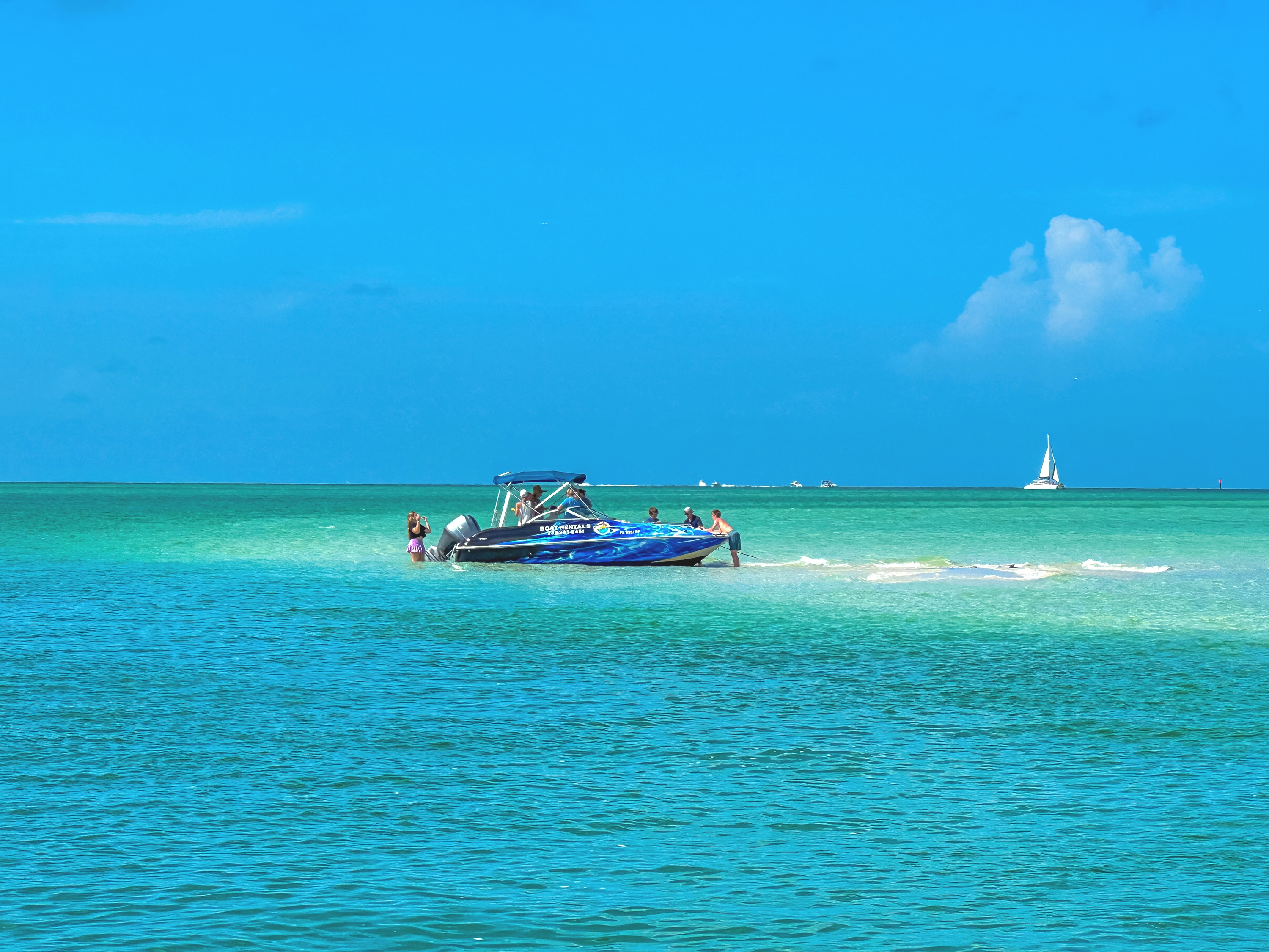 A motorboat with several people onboard in clear turquoise waters under a bright blue sky. A distant sailboat is visible on the horizon.