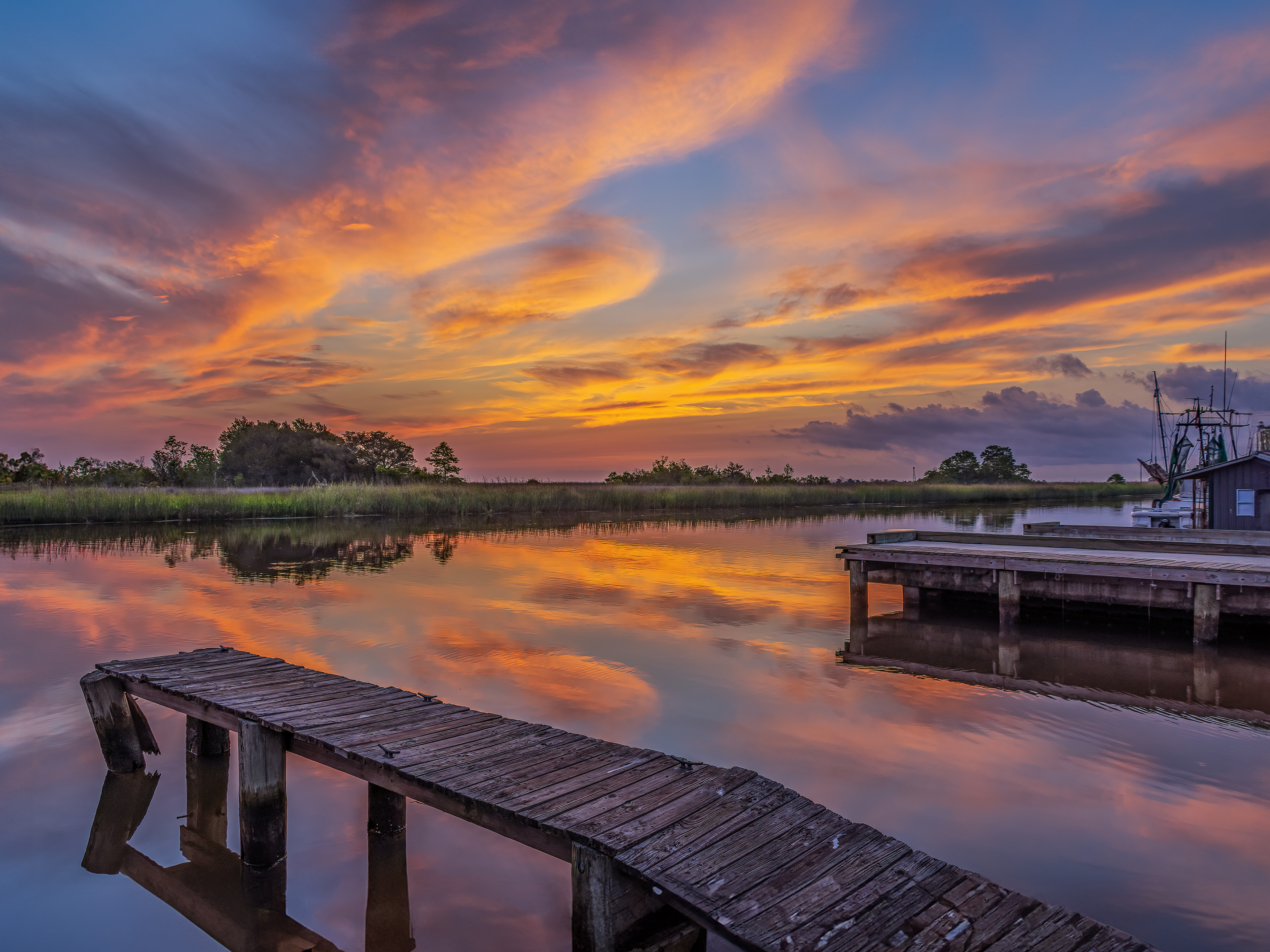 A sunset over a calm body of water reflects vibrant orange, yellow, and purple hues in the sky. Wooden docks extend into the water, and lush greenery lines the far shore.