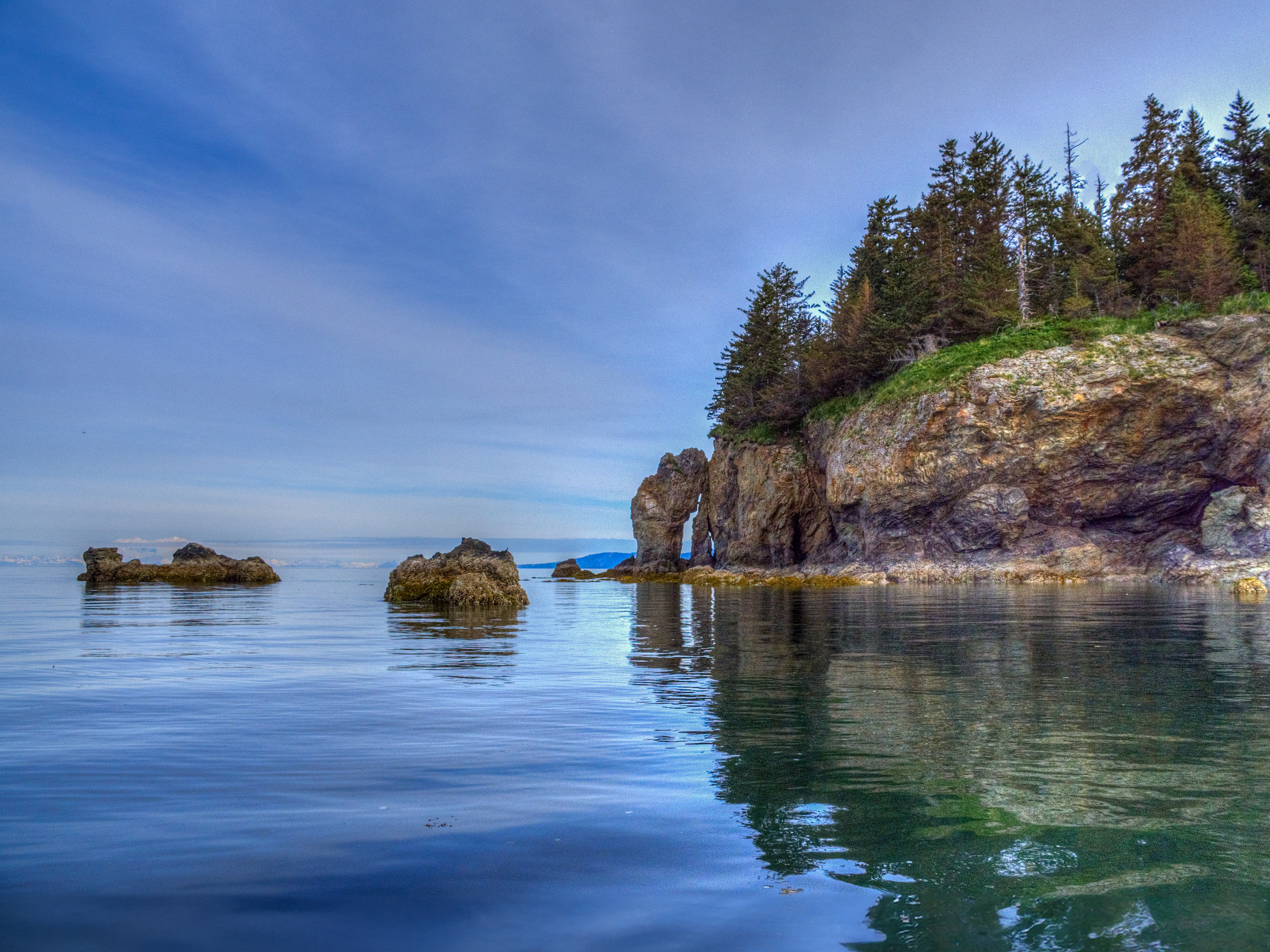 A calm, reflective body of water with rocky cliffs and evergreen trees on the right. The sky is clear and smaller rocks protrude from the water in the foreground.