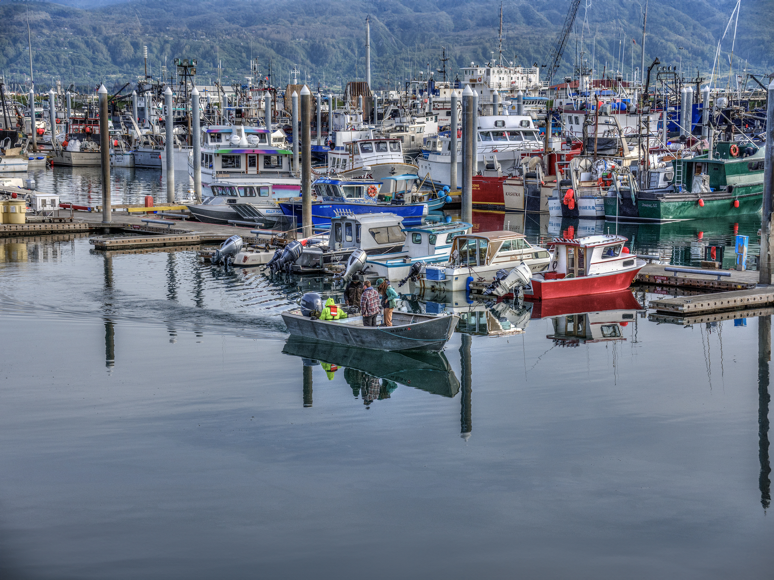 A marina filled with various boats and fishing vessels. A smaller boat with several people onboard is cruising through the calm waters in the foreground.