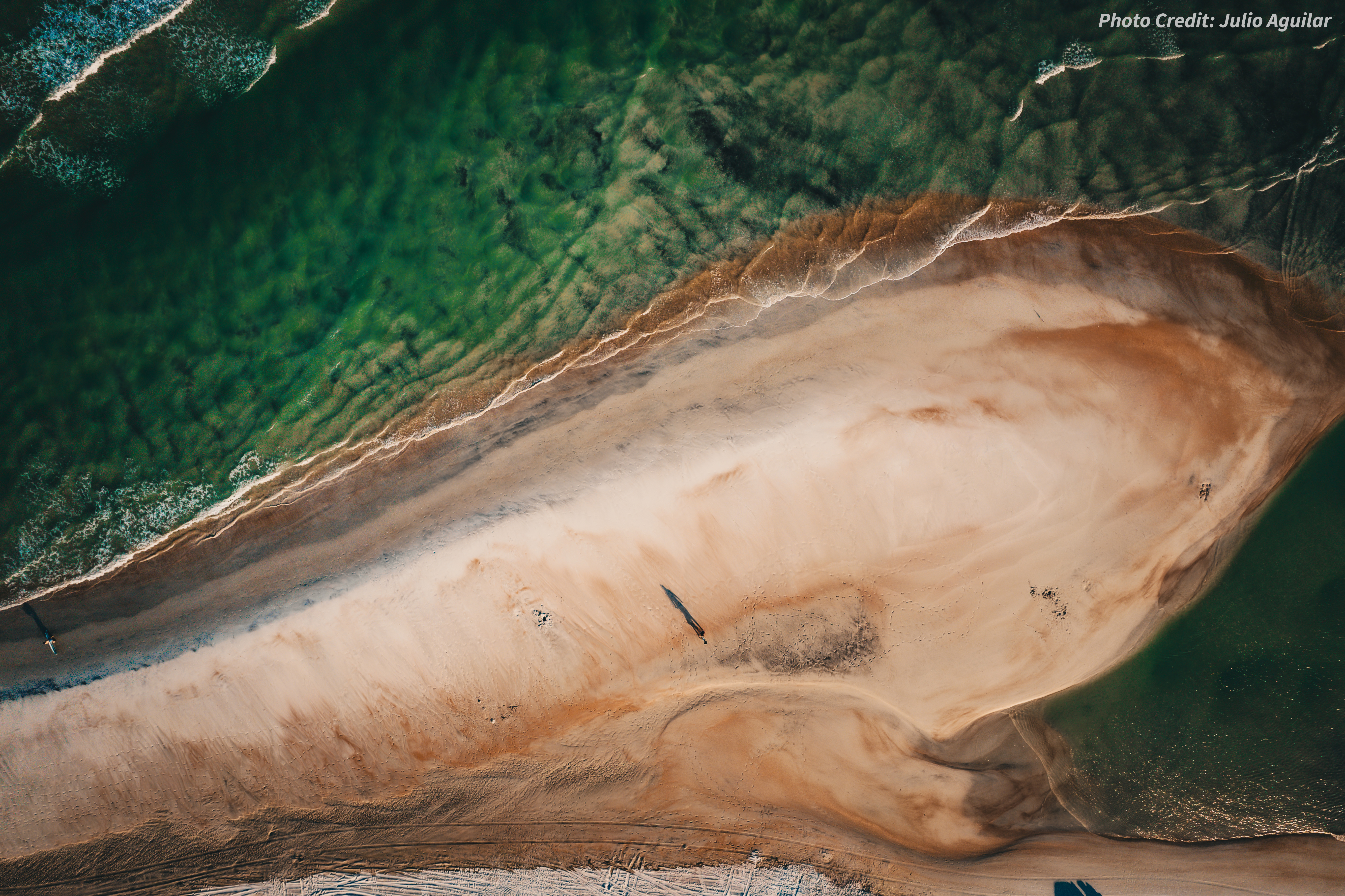 An aerial view of a sandbar going into green ocean waves as a person stands on the shore, casting a shadow far below.