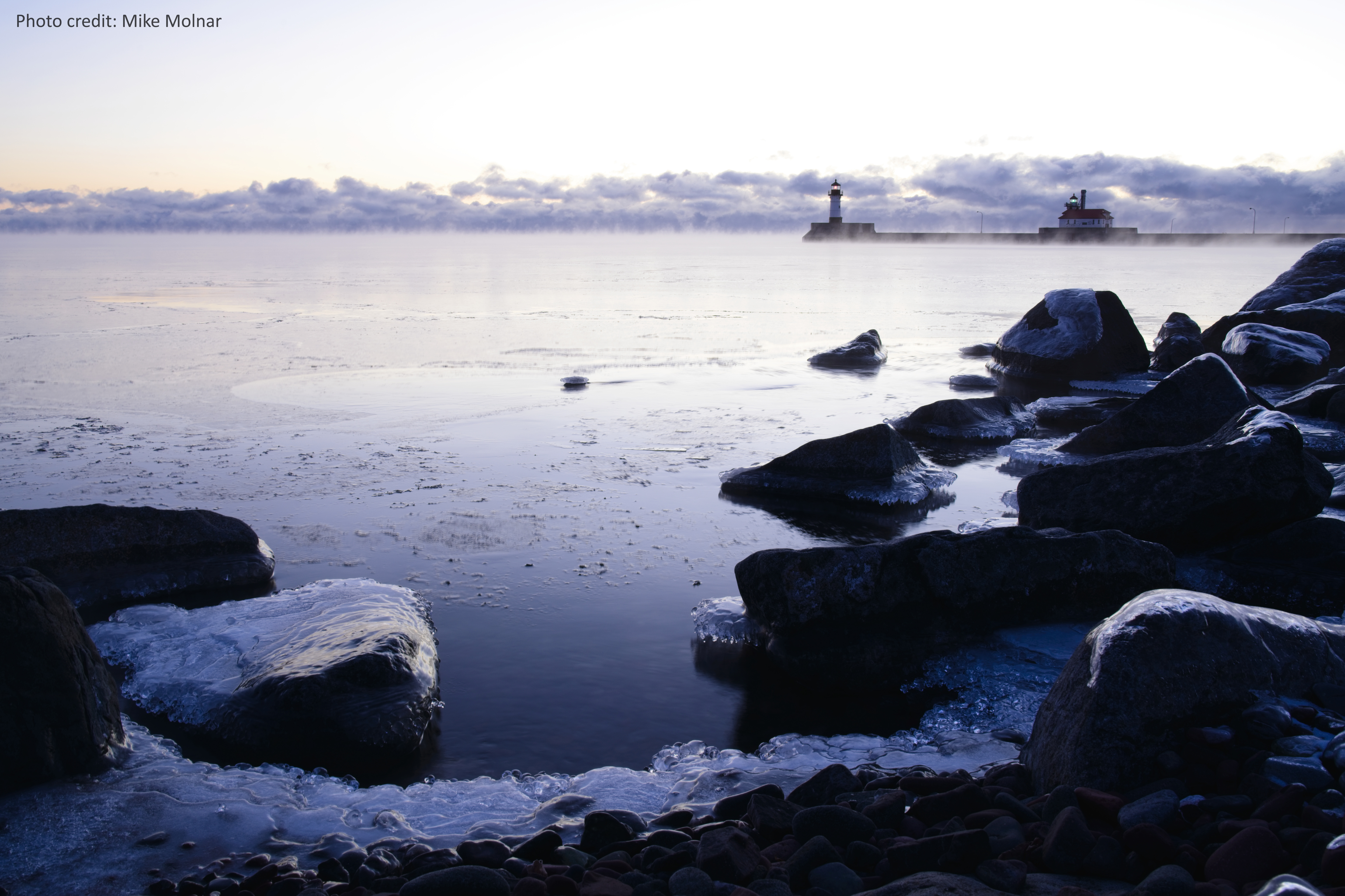 Fog and ice appear with rocks in the foreground and a lighthouse in the distance on Lake Superior.