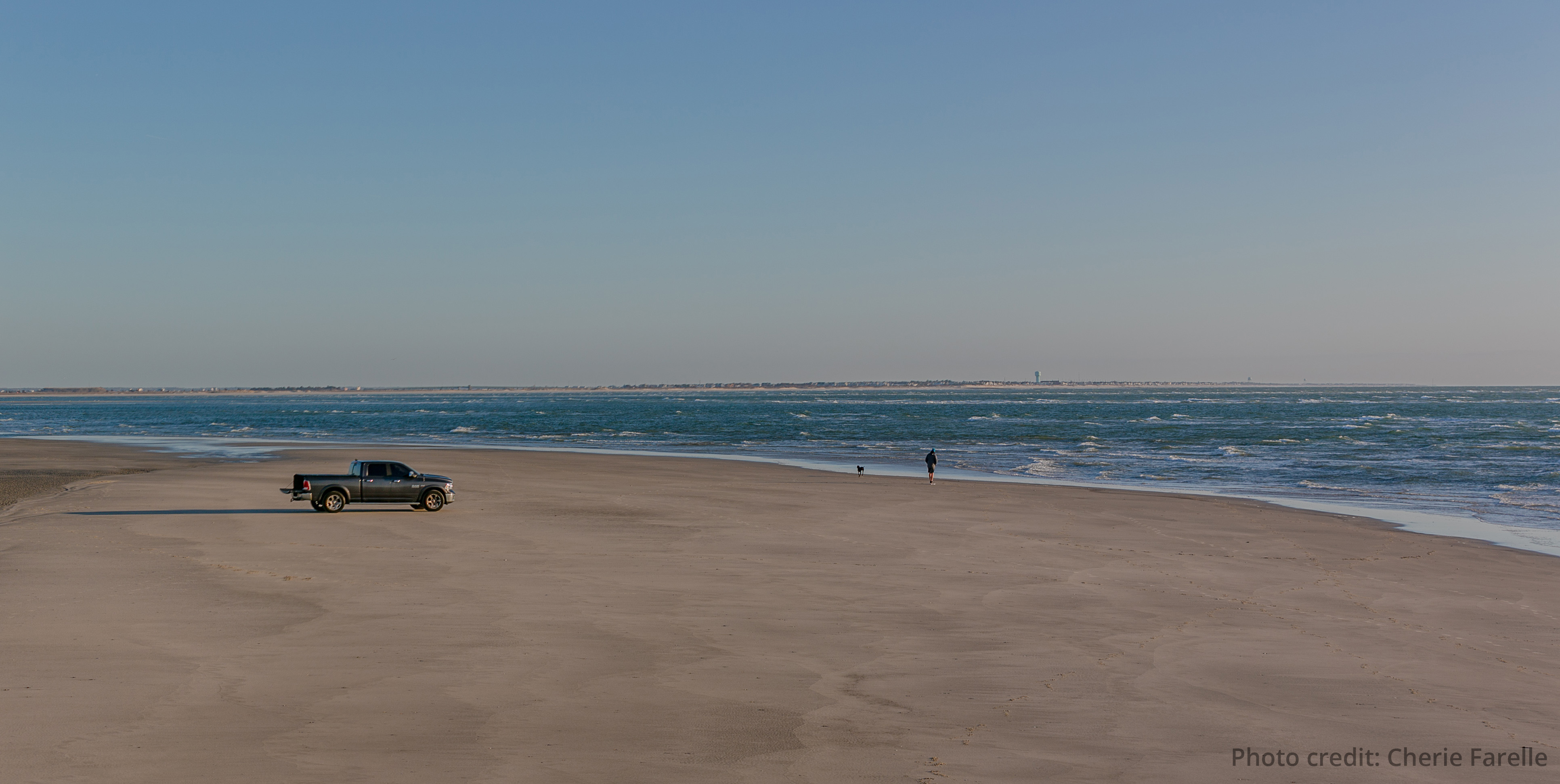 A truck sits parked on the beach as a person stands near the ocean waves.