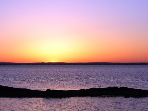 Sunrise over the water with land in the distance. A small strip of land is closer to the camera with water on either side.