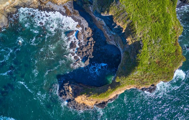 Aerial view of waves crashing on rocks and a hook-shaped cliff covered in green plants.