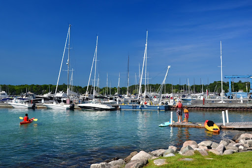 A marina full of boats. People stand on a pier near a person in a kayak in the water.
                