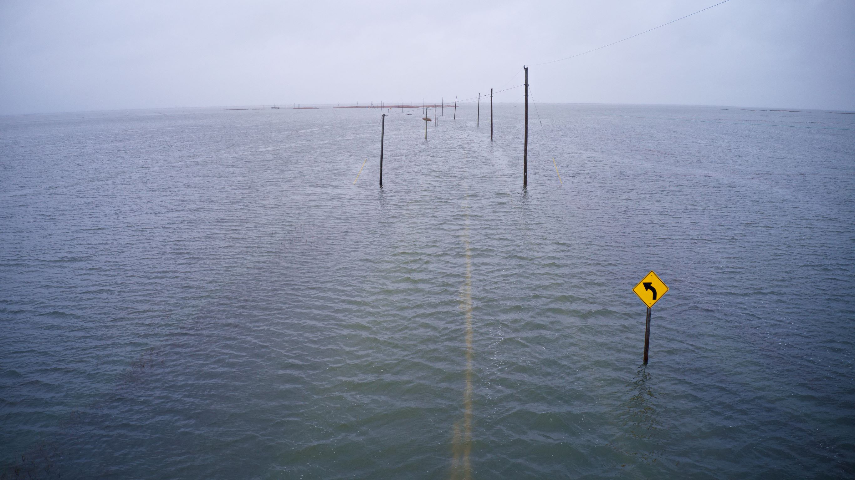: Image shows a roadway, turn sign, and electric cables flooded by water on all sides.