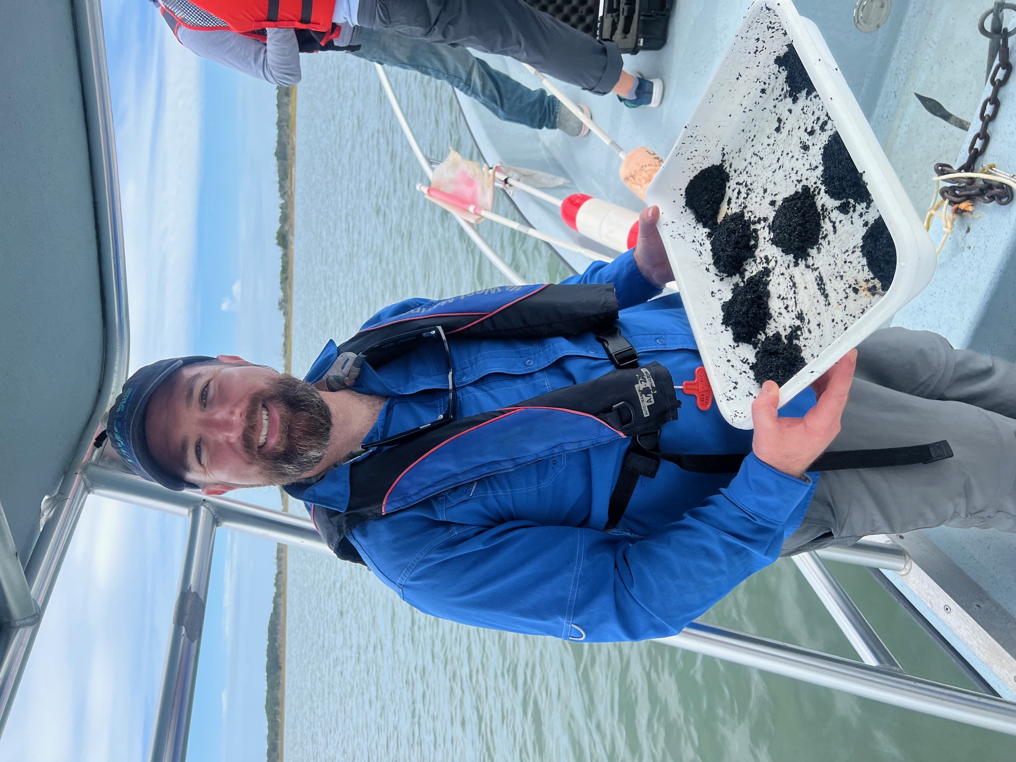 A person smiles holding a tray of dirt while on a boat.