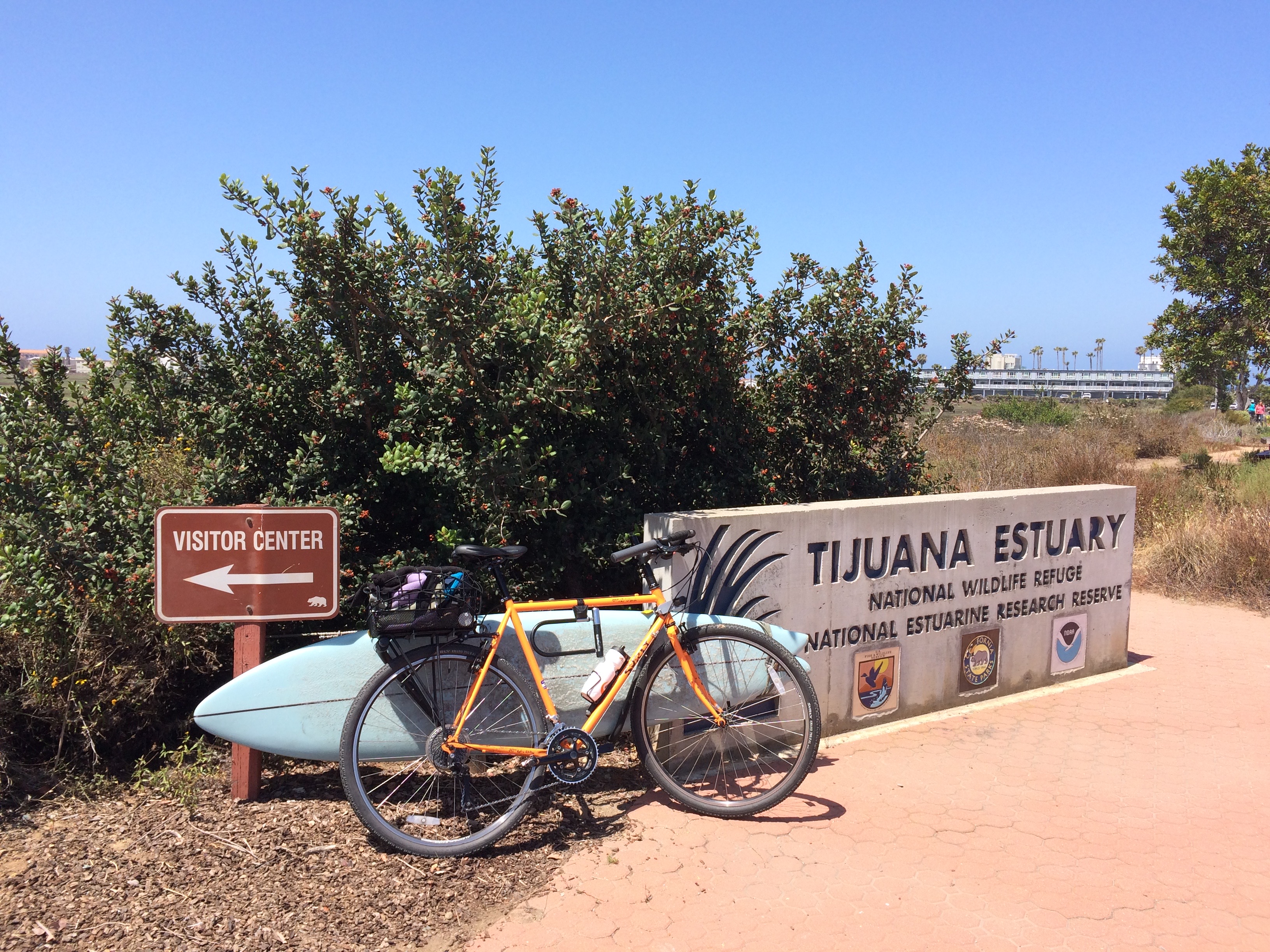 Along a marsh, a visitor center sign points left as a surfboard, a bike, and a Tijuana Estuary sign are seen.