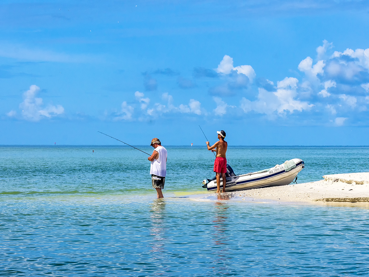 Two people cast fishing lines into ocean waters as they stand next to a sandy spit and a small boat.