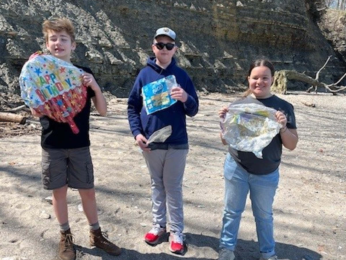 Three young students stand on a sandy beach holding up mylar balloons and other pieces of marine debris they collected.