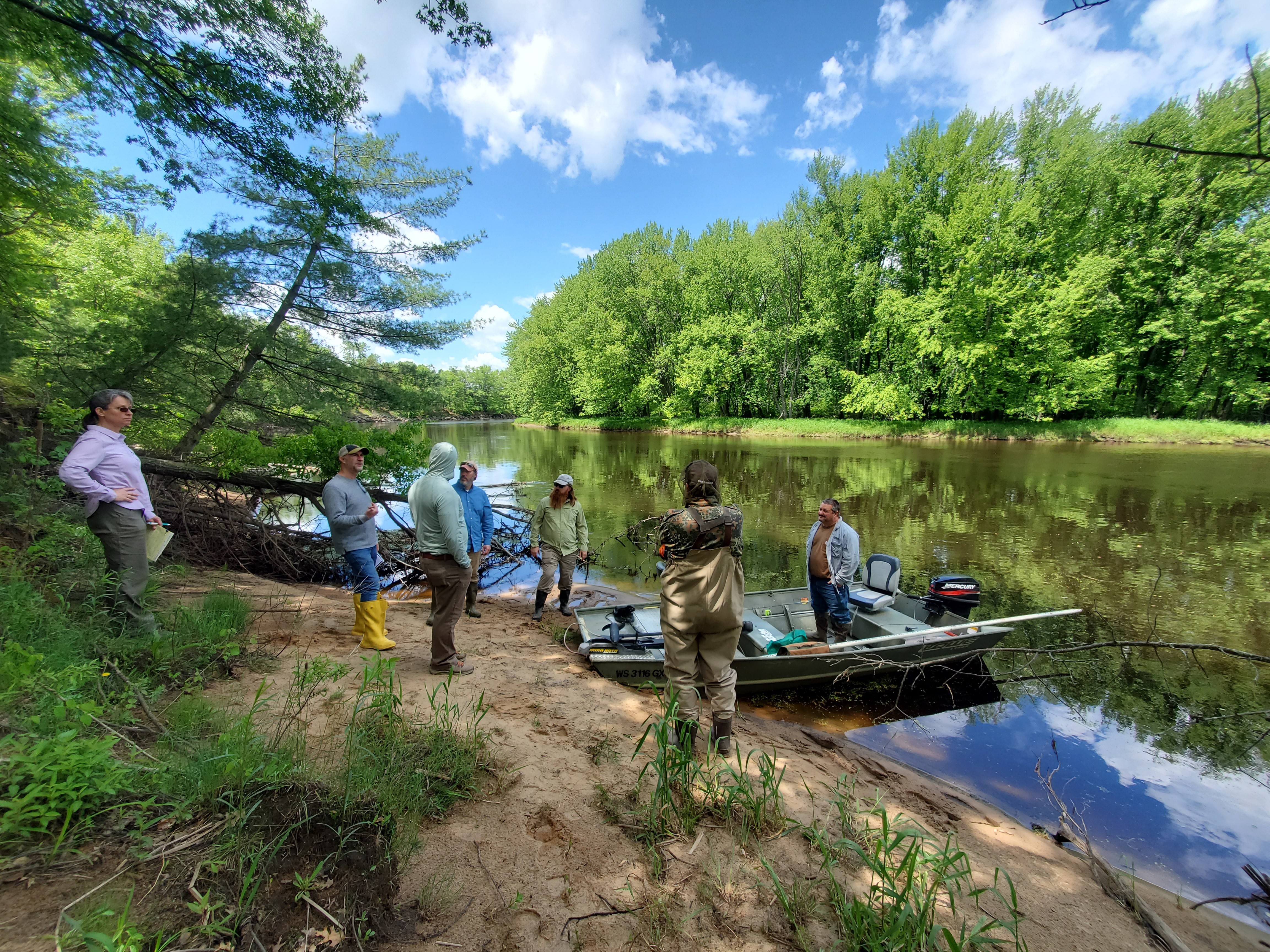 A group of people stand on a riverbank next to a boat. Trees are on either side of the water.