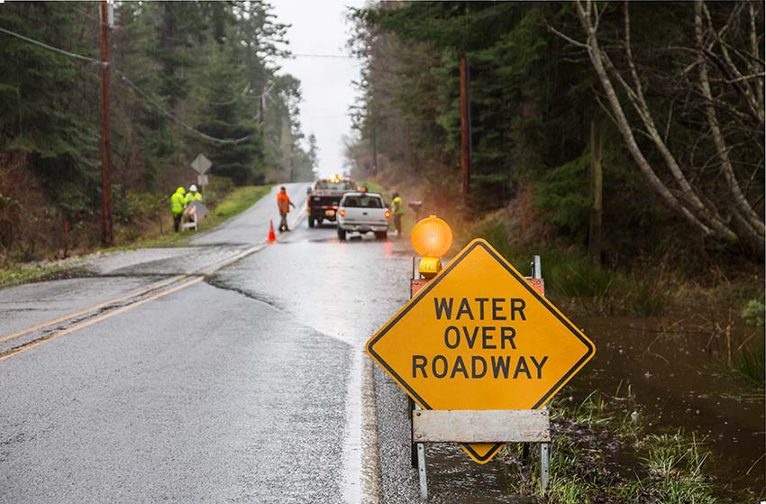 Flooded road with hazard warning sign and first responders assisting stranded car.